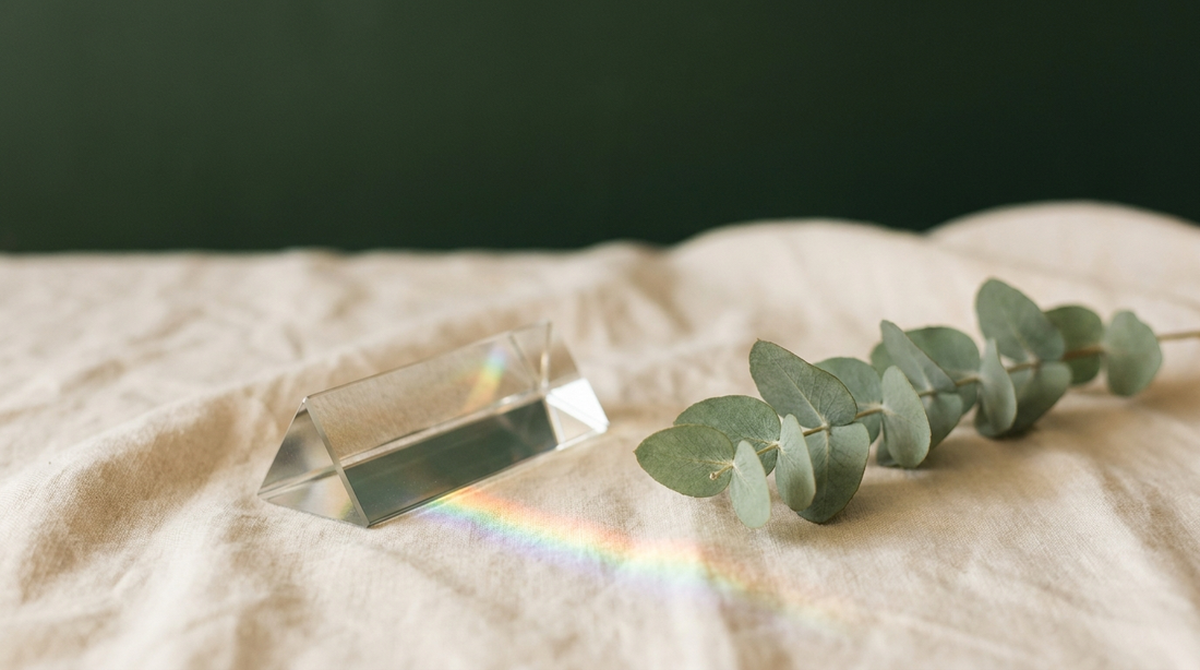 A glass prism on cream linen casting a soft faint rainbow of light, warm calm still-life.