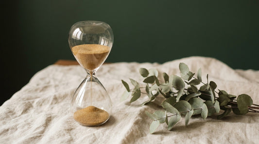 An elegant glass hourglass on cream linen in warm light, calm transition mood.