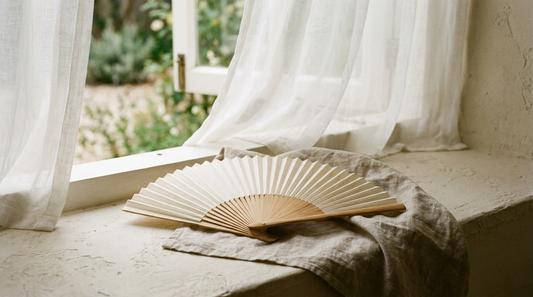 A folding hand fan and linen near an open window with airy curtains, soft natural light.