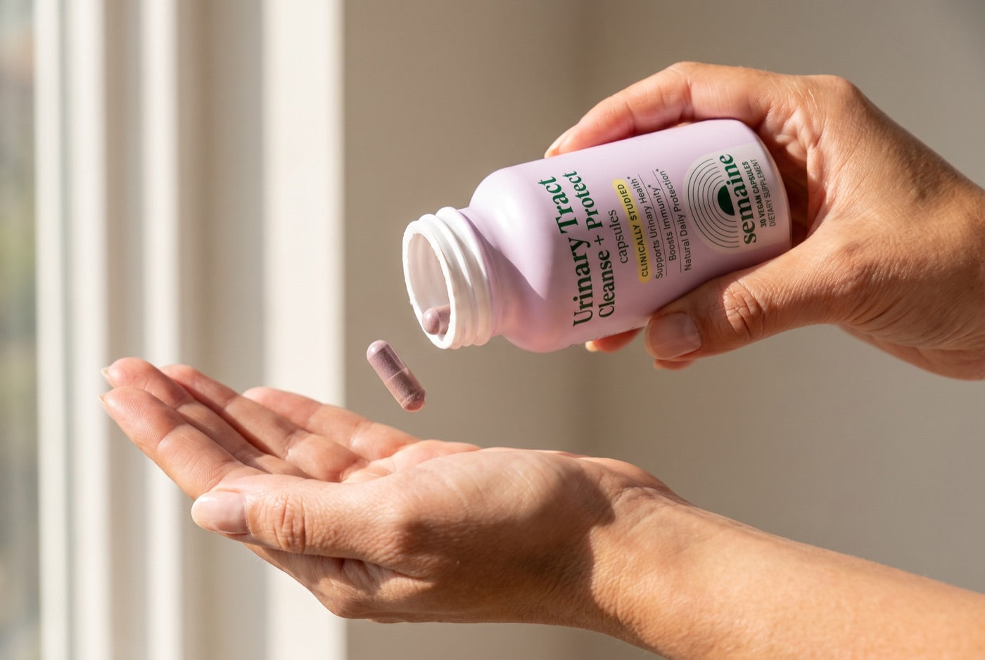 One cranberry-colored capsule falling from the Semaine Urinary Tract Cleanse + Protect bottle into a palm